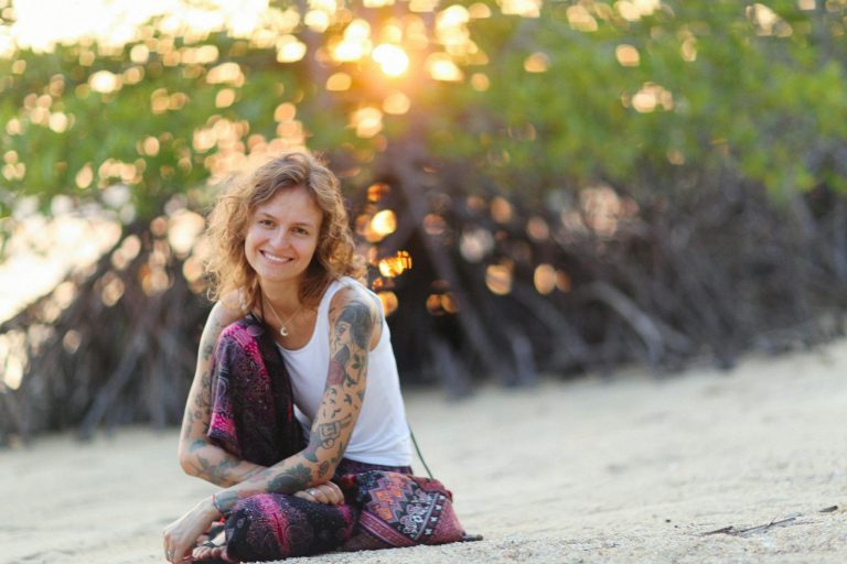 Caucasian woman with tattoos sitting on a sandy beach, enjoying the sunset.
