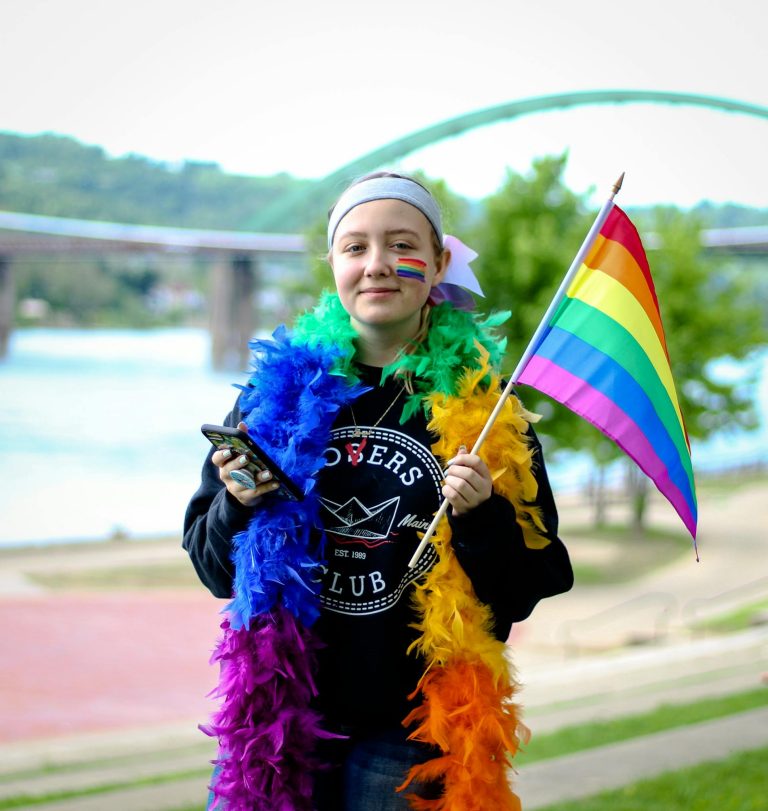 Young person joyfully celebrating Pride, holding a rainbow flag outdoors with colorful attire.
